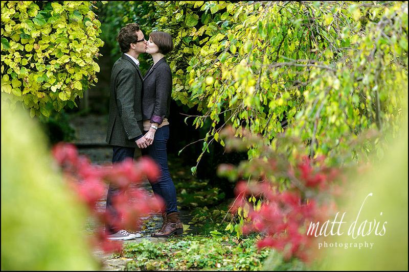 Vibrant autumnal colours make a great Engagement Photo of couple kissing. Taken in the gardens of Barnsley House Gloucestershire
